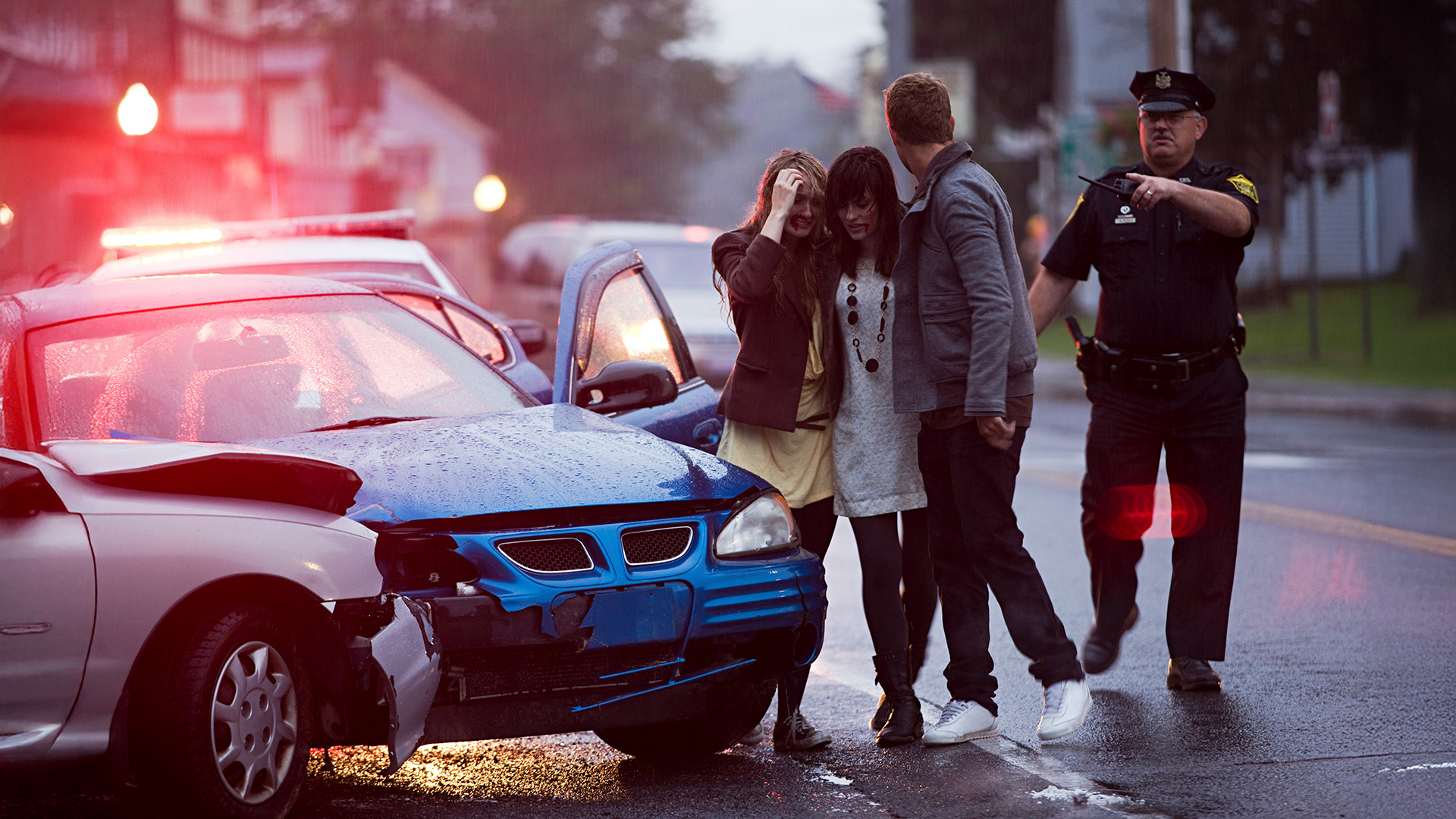 Two women, comforted by a man, stand beside a damaged blue car after an accident in the rain, while a police officer directs traffic nearby with flashing lights from a police vehicle.