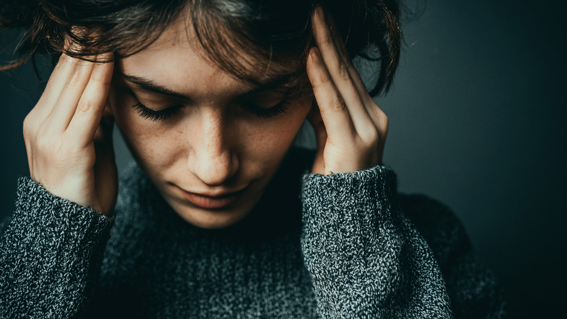 A woman wearing a sweater holds her head in her hands, indicating pain in her head.